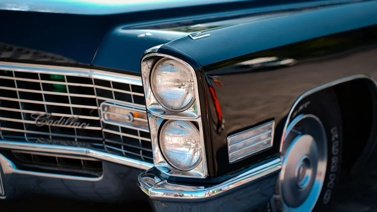 The front of a dark blue 1960s Cadillac, with a chrome grill and headlights, and a tire visible on the right side.